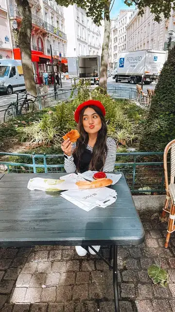 Camila de boina vermelha comendo um croissant numa mesa de café